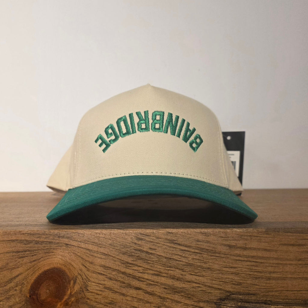 Beige and green cap with 'Bainbridge' text on a wooden surface