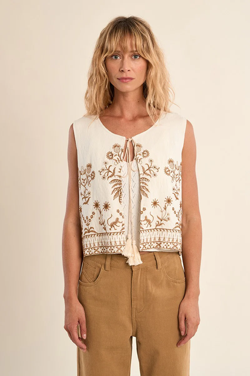 Woman wearing a sleeveless embroidered top and beige pants on a plain background