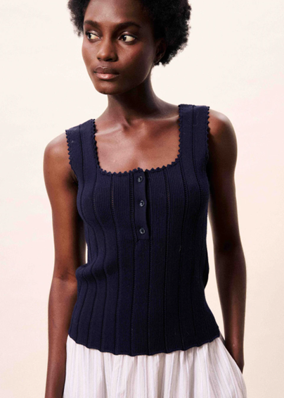 Woman wearing a navy blue sleeveless top with buttons on a light pink background