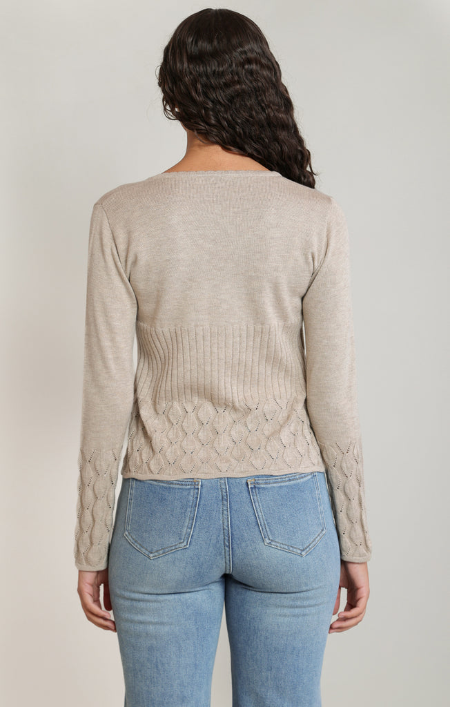 Person wearing a beige cable knit sweater and light blue jeans on a plain background