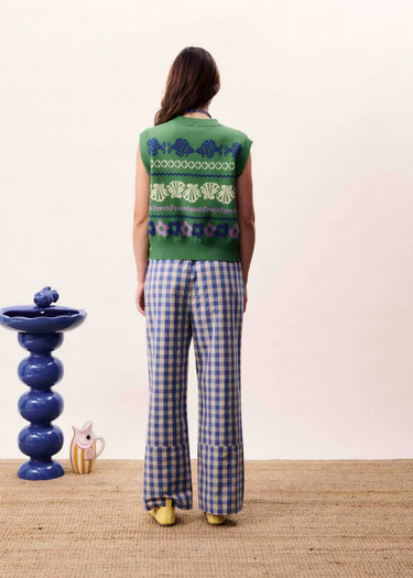 Person wearing a green patterned top and blue checkered pants standing in front of a white wall with a decorative blue vase and small figurine on a table.