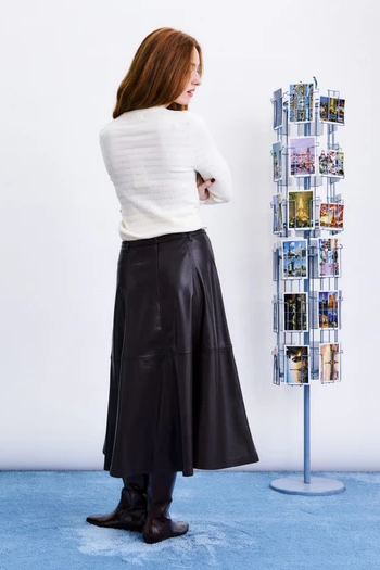 woman with crossed arms from behind in a cream top and maroon midi skirt standing next to postcard stand