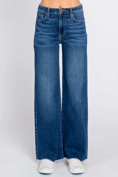 A person wearing high-waisted, wide-leg blue denim jeans with a raw frayed hem, paired with white sneakers, shown from the waist down on a light gray studio background.