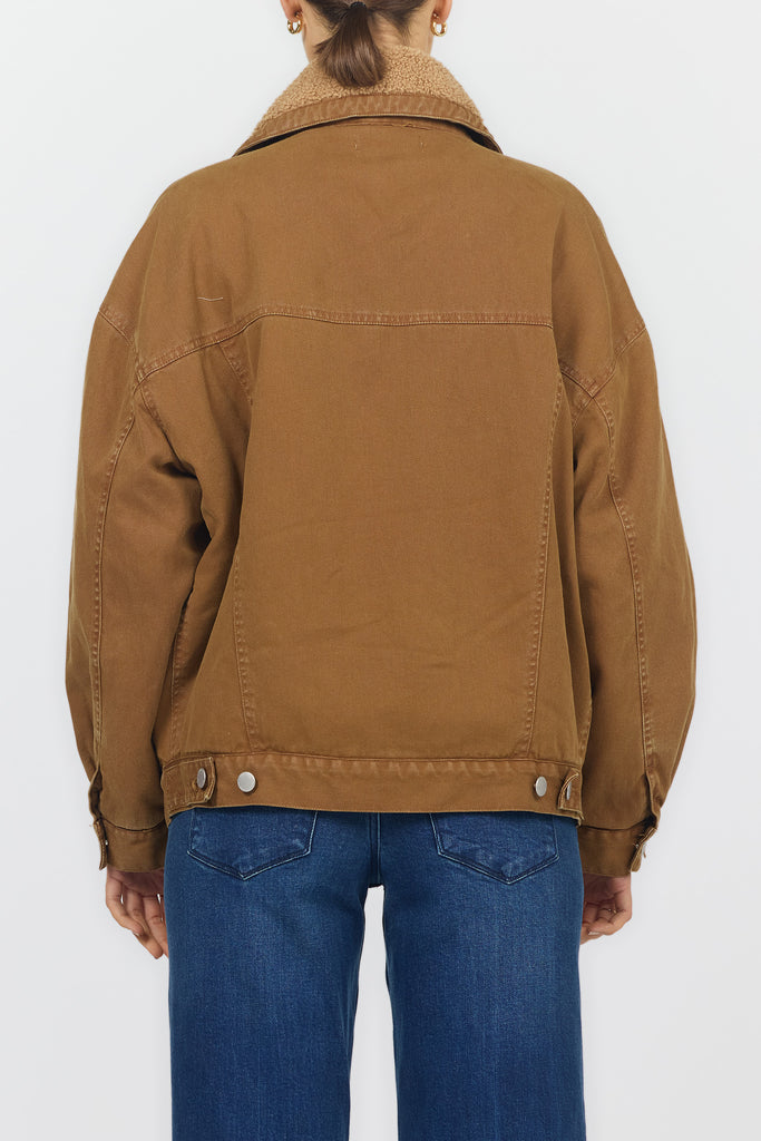 A person viewed from the back wearing a tan-brown jacket with a light beige sherpa collar and blue jeans, against a plain light gray studio background.