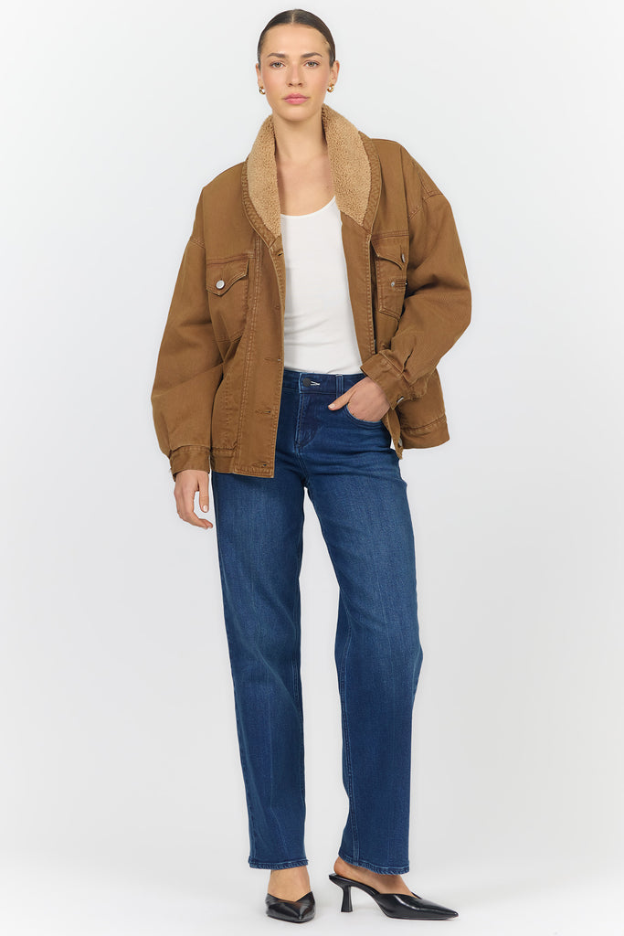 A model stands against a light gray studio background wearing a brown jacket with a tan sherpa collar, a white top, blue jeans, and black pointed mule heels.