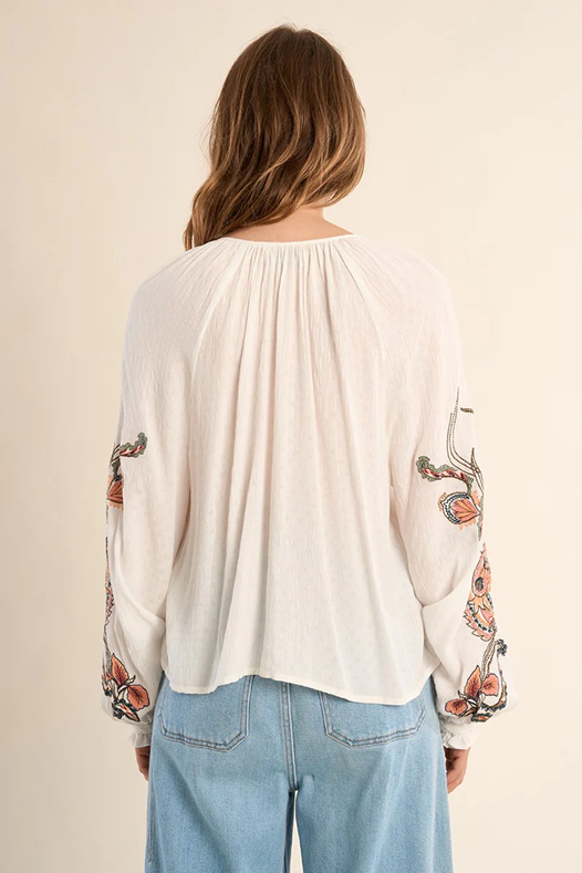 Woman wearing a white blouse with floral embroidery on sleeves against a beige background