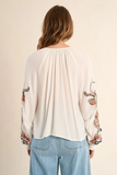 Woman wearing a white blouse with floral embroidery on sleeves against a beige background