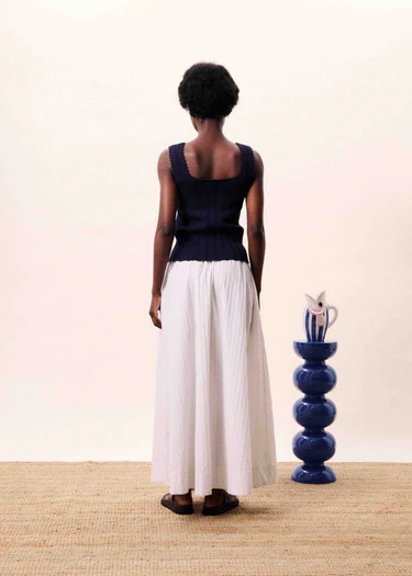 Person wearing a black sleeveless top and white pants standing in front of a blue vase on a beige surface.
