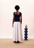 Person wearing a black sleeveless top and white pants standing in front of a blue vase on a beige surface.
