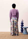 Person wearing a purple sweater and patterned pants standing in a room with a table and decorative items.