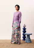 Woman wearing a purple sweater and floral pants standing in a room with a blue stool and decorative items.