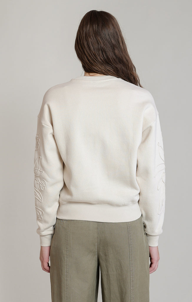 A woman viewed from the back wearing a cream-colored crewneck sweatshirt with tonal embroidered details on the sleeves, paired with olive green trousers, against a neutral light gray background.