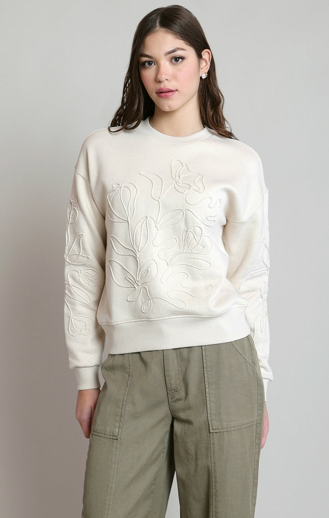 A woman modeling a cream-colored crewneck sweatshirt with tonal floral embroidery on the front and sleeves, paired with olive green cargo pants, against a neutral gray background.