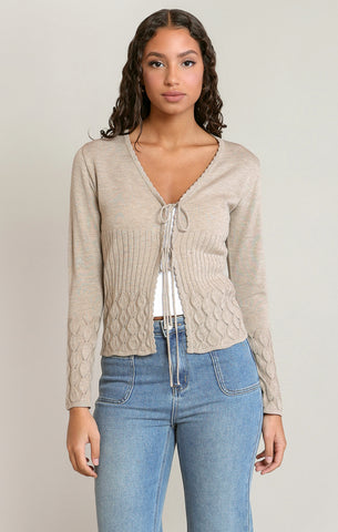 Woman wearing a beige cable knit cardigan and blue jeans on a plain background