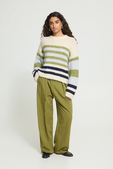 woman in horizontal striped sweater and green pants with one hand in a pocket against white background
