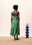 Woman wearing a green dress with a white pattern, standing in a room with a blue vase on a table.