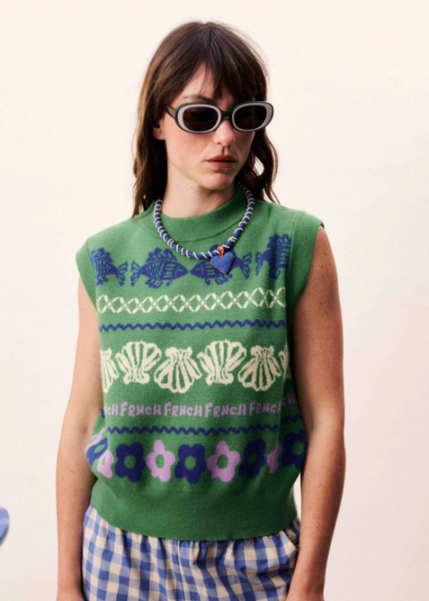Woman wearing a green sleeveless sweater with patterns and text, sunglasses, and a necklace.