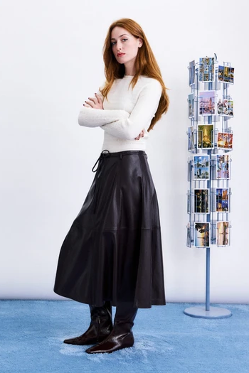 woman with crossed arms in a cream top and maroon midi skirt standing next to postcard stand