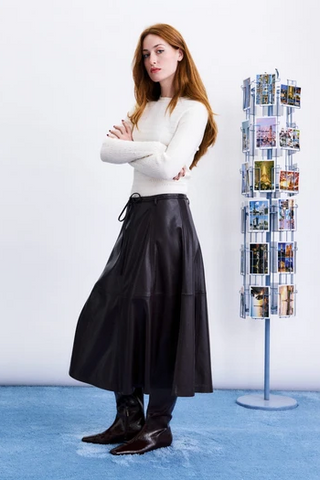 woman with crossed arms in a cream top and maroon midi skirt standing next to postcard stand