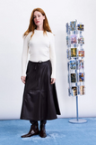 woman with crossed arms in a cream top and maroon midi skirt standing next to postcard stand