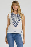 Woman wearing a white sleeveless top with blue floral embroidery and blue jeans on a light gray background