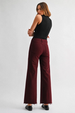 A woman is shown from the back wearing high-waisted, wide-leg burgundy pants with a frayed hem, a black sleeveless top, and black shoes, set against a plain light gray studio background.
