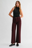 A woman modeling a pair of high-waisted, wide-leg maroon jeans with frayed hems, paired with a black sleeveless top and black loafers, against a light gray studio background.