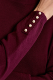 Close-up of a burgundy knit sweater sleeve featuring a cuff pocket with five white pearl buttons, worn by a model.