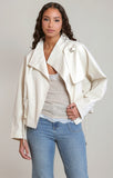 A woman modeling a cream-colored moto jacket with a unique draped collar and a decorative silver-toned brooch on the lapel, styled over a light gray top and blue jeans against a neutral studio background.